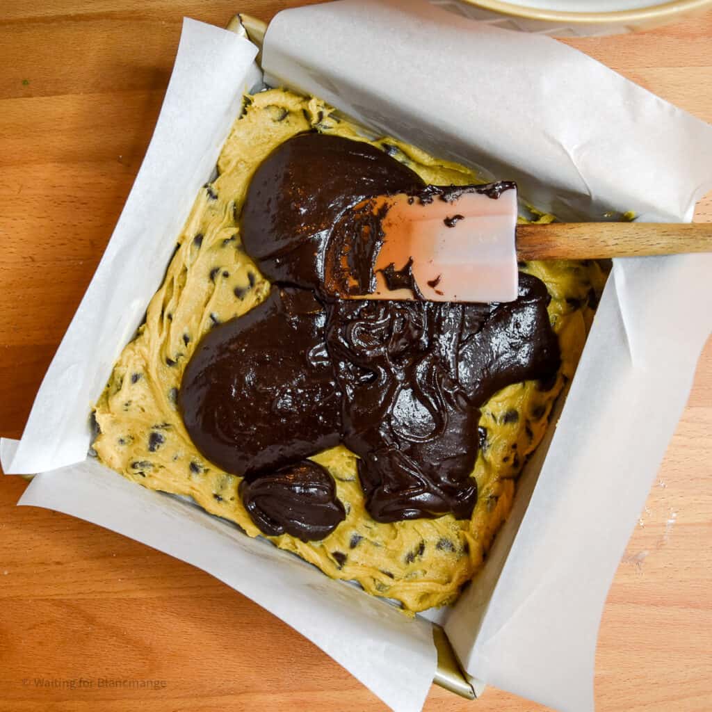 Adding fudgy brownie batter over top chocolate chip cookie dough in a lined square pan.