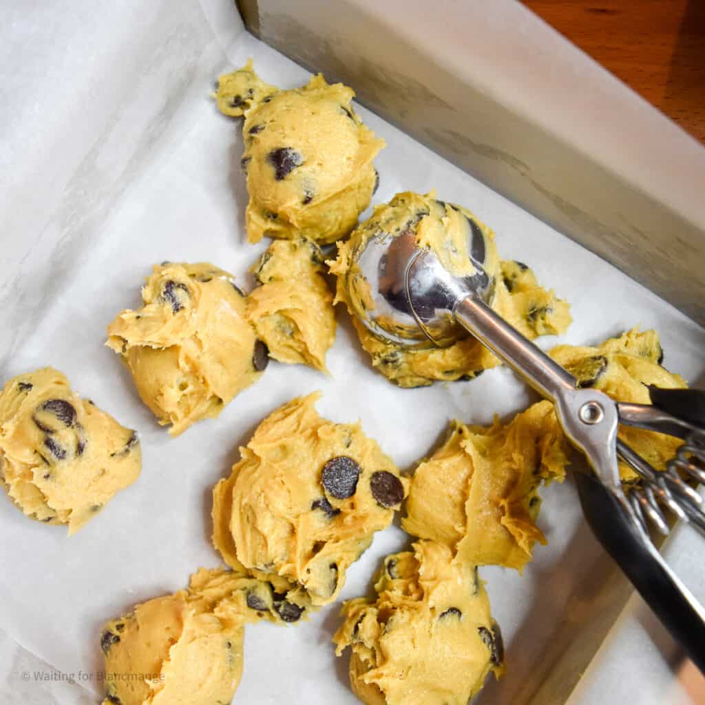 Adding scoops of cookies dough to lined pan to spread into a layer.