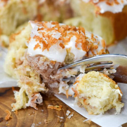 A fork cutting into a soft coconut cinnamon roll topped with cream cheese and toasted coconut on a wooden board.