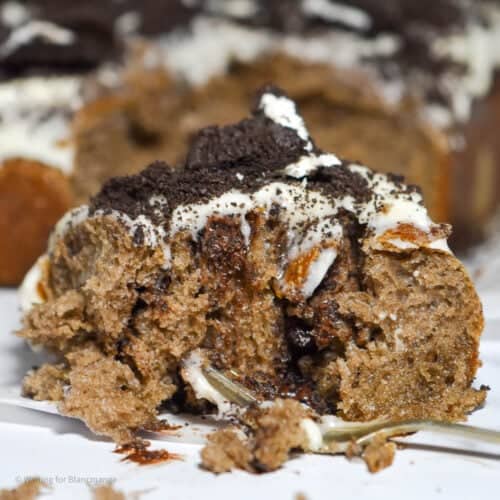 Close up image of a gooey Cookies and Cream Cinnamon Roll with cream cheese frosting and Oreo crumbs on top. The roll has been half eaten, exposing the insides layers of chocolate and Oreo filling. There is a small dessert fork is in front of the roll.