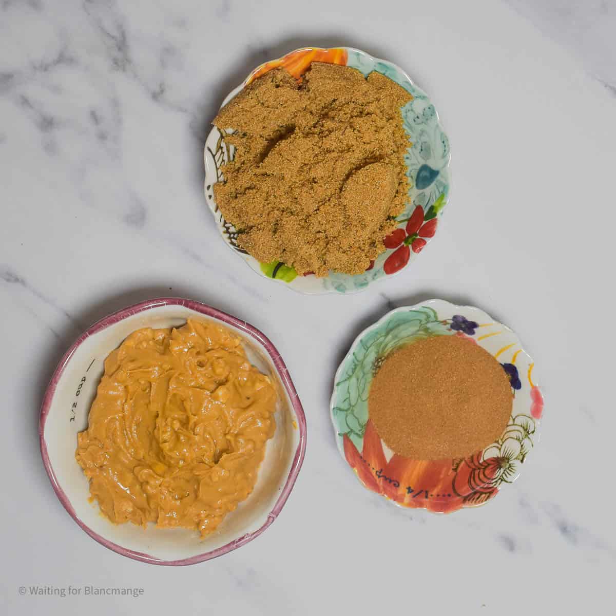 Overhead shot of three small bowls, one holding crunchy peanut butter, another holding light brown sugar and another holding ground cinnamon. The bowls are sat on a white marble counter top.