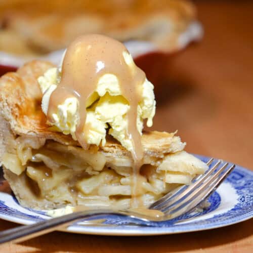 A slice of homemade apple pie on a blue and white plate with a dessert fork. The pie is topped with ice cream and a generous drizzle of caramel sauce.