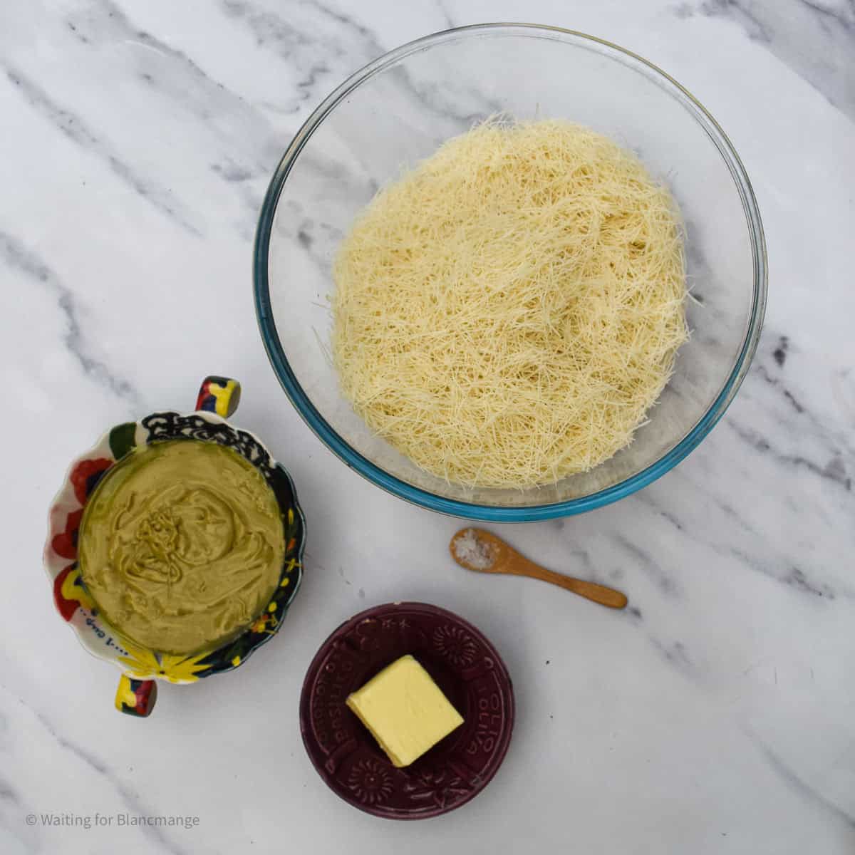 Overhead shot of bowls holding the ingredients for making Dubai-style pistachio filling including pistachio paste, butter, kataifi pastry and salt.